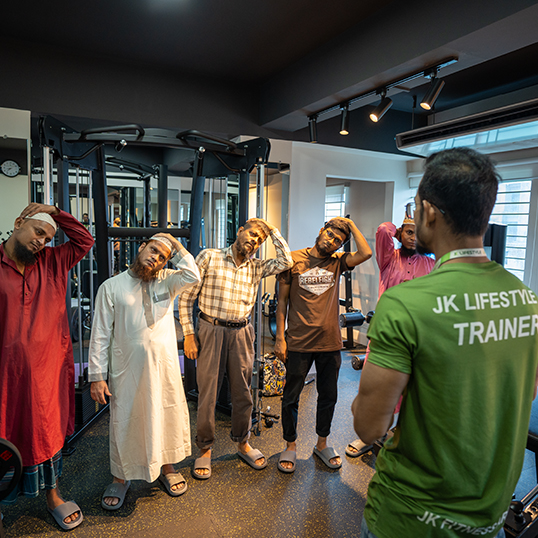 Group of men, including some in traditional attire, performing neck stretches under the guidance of a trainer in a modern gym, symbolizing building a fitness culture.